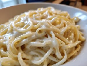 Close-up of spaghetti noodles generously coated in a rich, creamy vegan alfredo sauce, showing flecks of pepper.