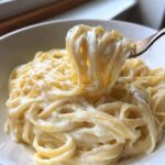 Close-up of fettuccine noodles coated in a rich, creamy vegan alfredo sauce being lifted by a fork.