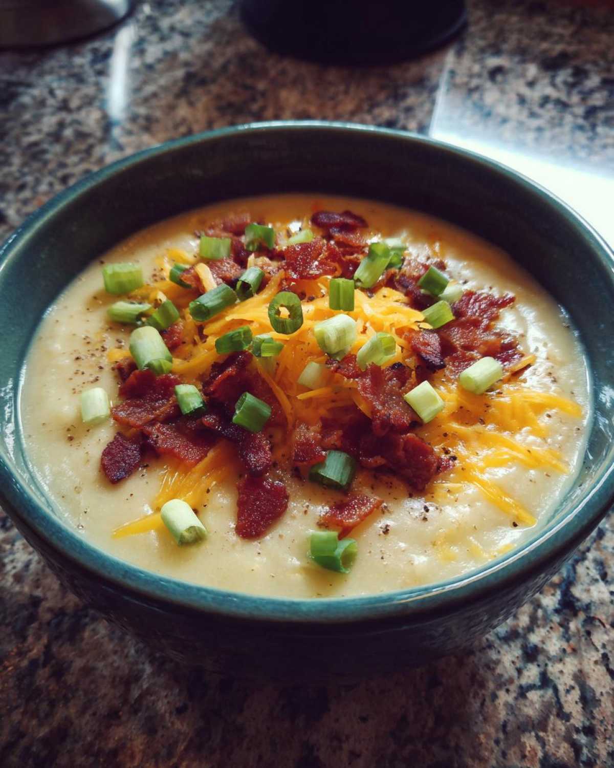 A close-up of creamy potato soup recipe topped generously with shredded cheddar cheese, crispy bacon bits, and fresh green onions.