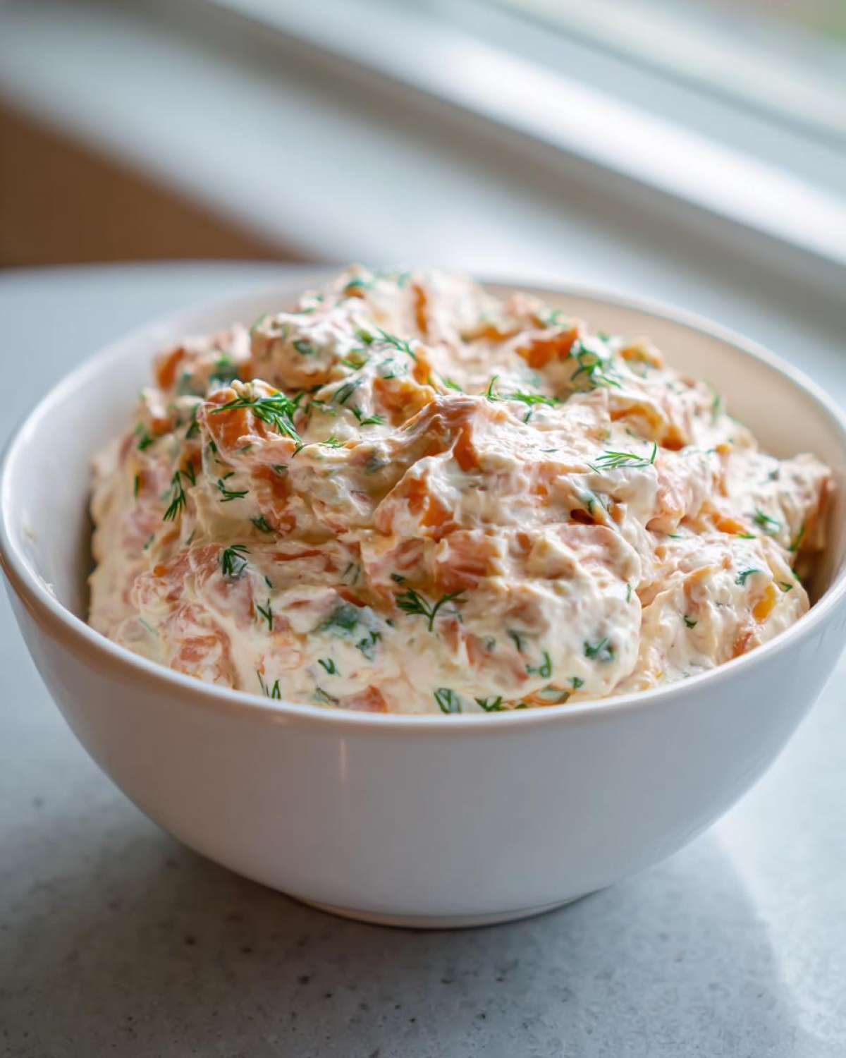 A white bowl filled with creamy, chunky smoked salmon dip garnished generously with fresh dill.
