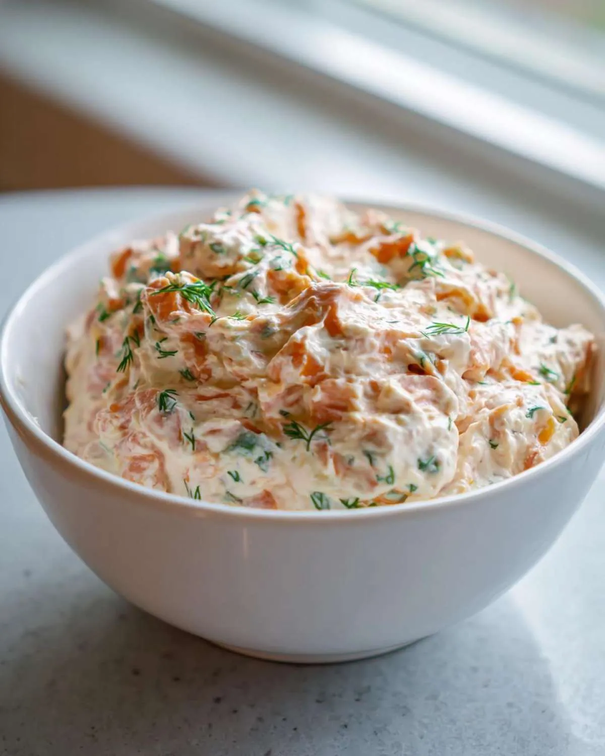 A white bowl filled with creamy, chunky smoked salmon dip garnished generously with fresh dill.