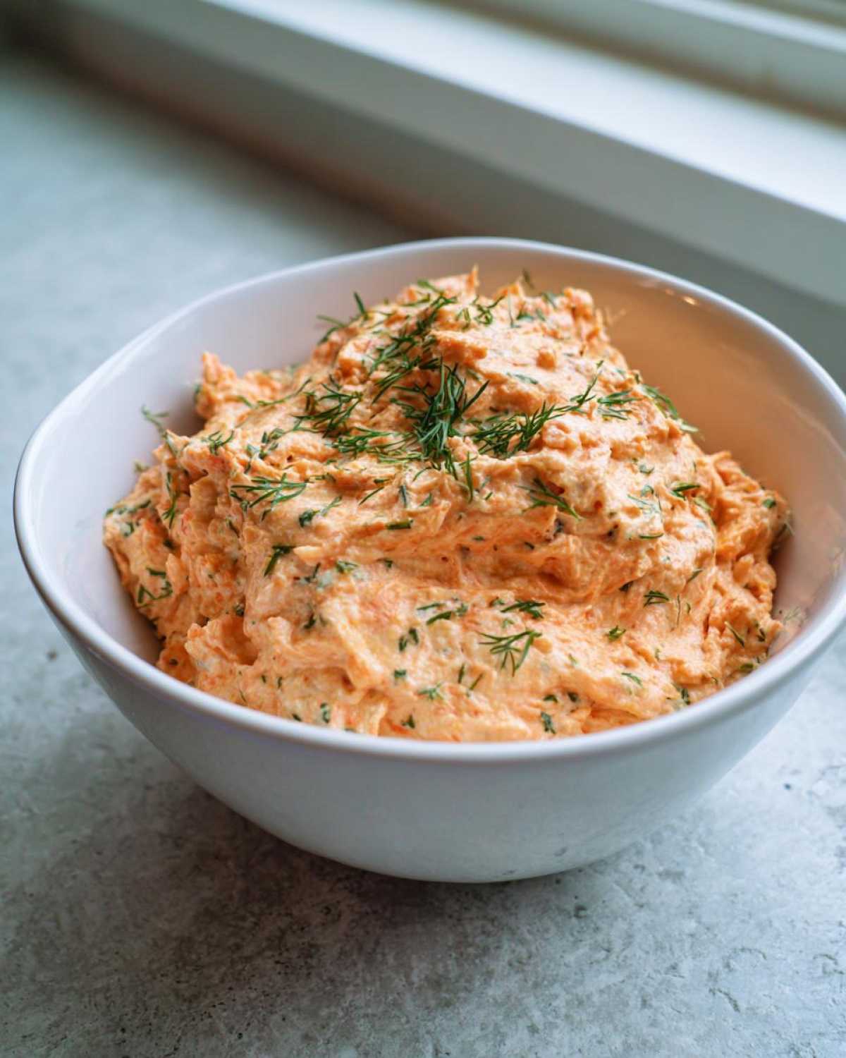 A bowl filled with creamy, orange-hued smoked salmon dip, garnished generously with fresh dill.