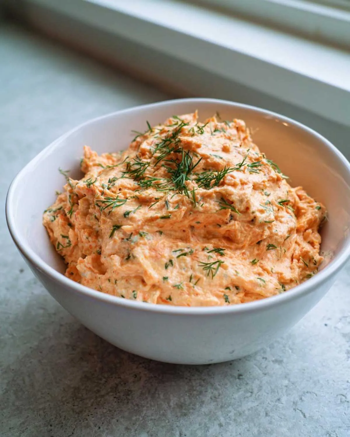 A bowl filled with creamy, orange-hued smoked salmon dip, garnished generously with fresh dill.