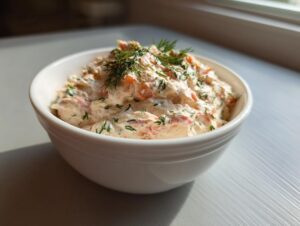 A creamy, pinkish smoked salmon dip garnished with fresh dill, served in a white bowl near a window.