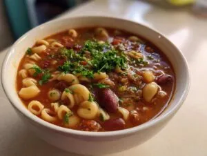Close-up of a white bowl filled with rich, tomato-based pasta fagioli soup, topped with fresh parsley.