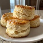 Close-up of four flaky buttermilk biscuits stacked on a white plate with a light glaze.