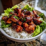 Close-up of a bowl filled with white rice, topped with glazed, dark-sauced harissa chicken pieces and fresh green lettuce.