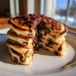 A tall stack of fluffy chocolate chip pancakes, cut to show the interior, drizzled with melted chocolate.