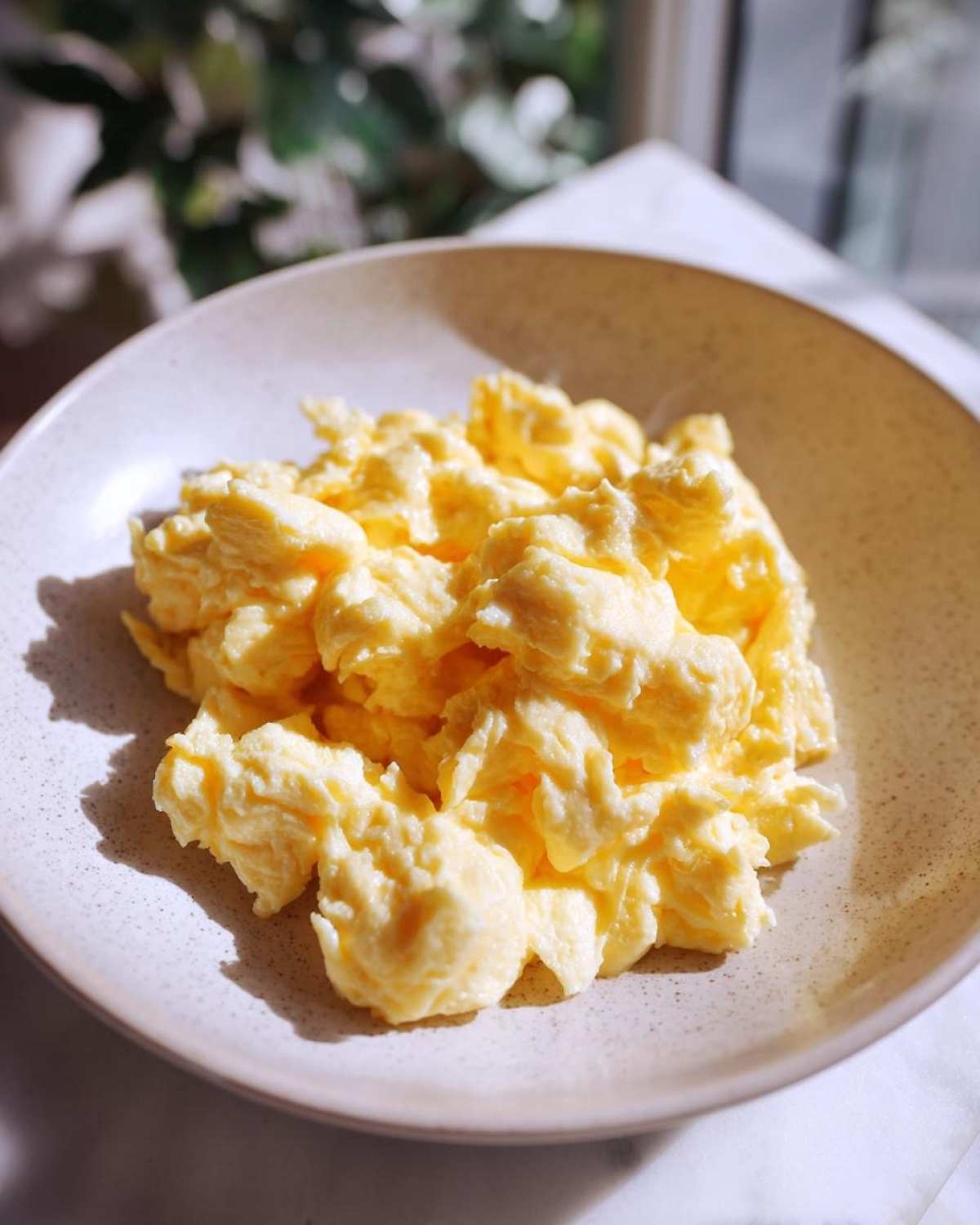 Close-up of perfectly fluffy scrambled eggs served in a light, speckled ceramic bowl.