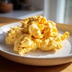 A close-up of perfectly fluffy scrambled eggs served on a light, speckled ceramic plate.