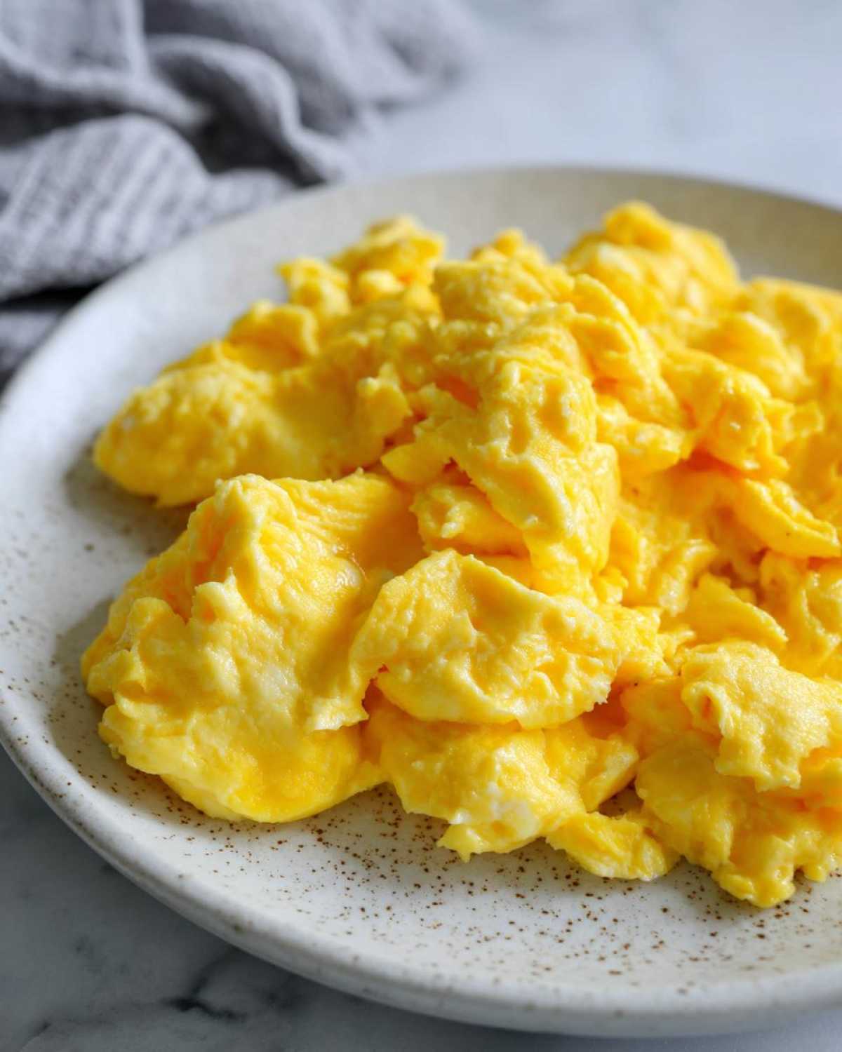 Close-up of bright yellow, fluffy scrambled eggs served on a speckled ceramic plate.