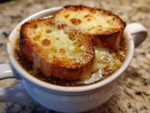 Close-up of a white crock filled with rich french onion soup topped with cheesy, broiled croutons.