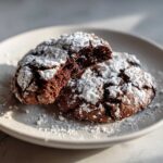 Two rich, dark chocolate crinkle cookies dusted heavily with powdered sugar on a light plate.