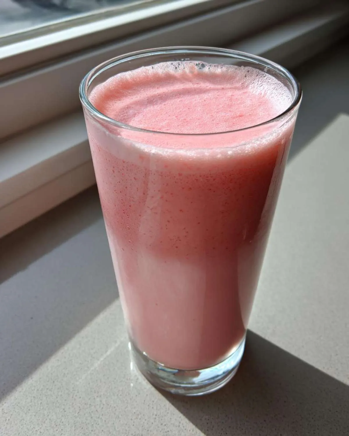 A tall glass filled with frothy, pink homemade strawberry milk sitting on a light countertop near a window.