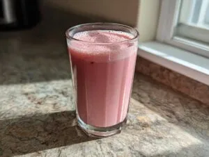 A tall glass filled with frothy pink strawberry milk sitting on a speckled countertop near a window.