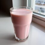 A tall glass filled with frothy, pink strawberry milk sitting on a bright white windowsill.