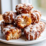 Close-up of a stack of golden brown apple fritter bites drizzled with white glaze, one has a bite missing.