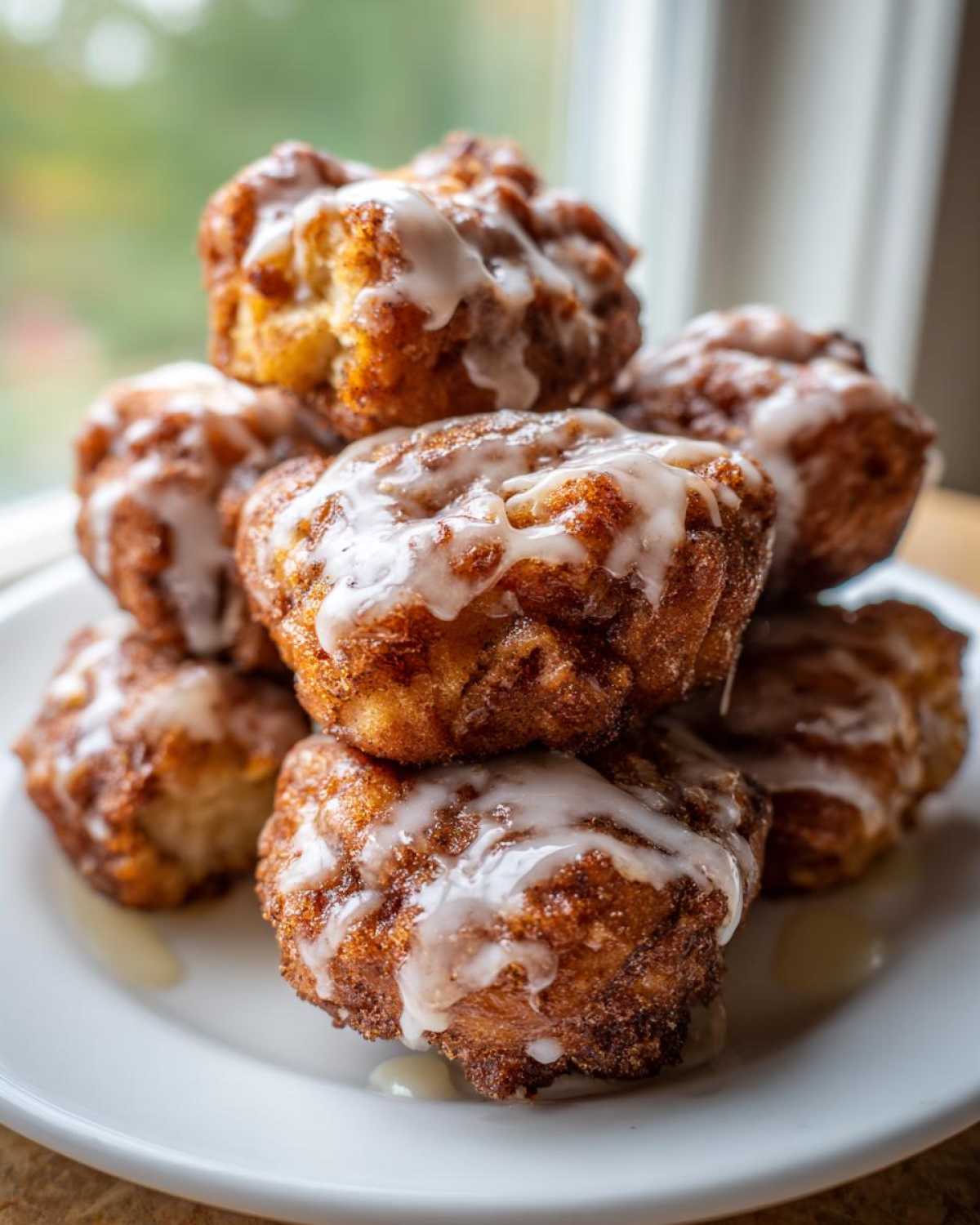 A stack of golden brown, deep-fried apple fritter bites drizzled with white glaze, served on a white plate.
