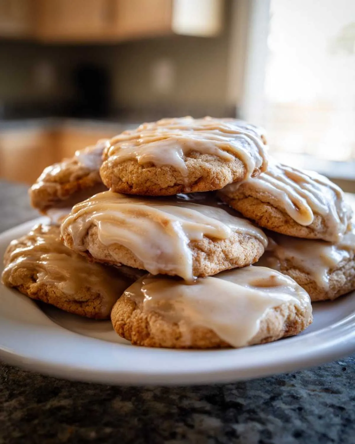 A stack of soft, glazed maple cookies piled high on a white plate in a kitchen setting.