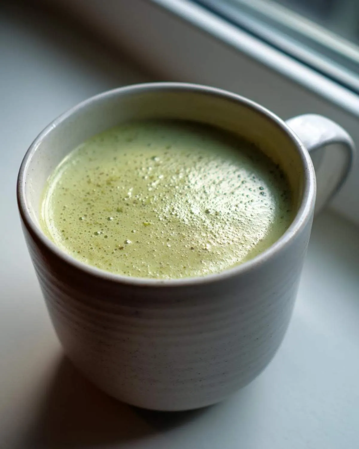 Close-up of a light green, frothy beverage in a textured white mug, which is not the celery soup.