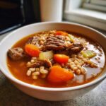 Close-up of a steaming bowl of rich beef barley soup featuring chunks of tender beef, bright carrots, and barley.