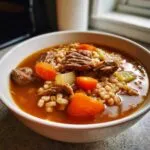 Close-up of a steaming bowl of rich beef barley soup featuring chunks of tender beef, bright carrots, and barley.