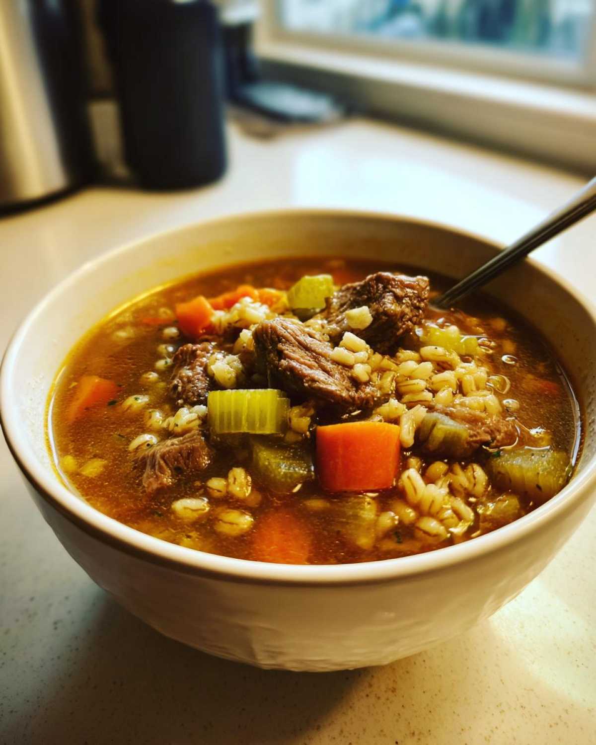A close-up of a steaming bowl of rich beef barley soup containing chunks of beef, carrots, celery, and barley.
