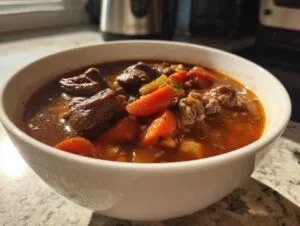 A close-up of a white bowl filled with rich, dark beef barley soup, featuring chunks of beef and bright orange carrots.