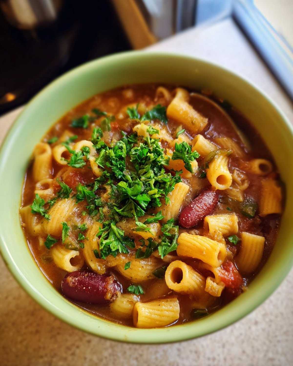 Close-up of a green bowl filled with rich pasta fagioli soup, featuring tubular pasta, kidney beans, and fresh parsley garnish.