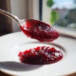 A spoonful of glistening, deep red homemade raspberry jam is held above a dollop on a white plate.