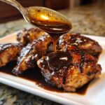 A spoonful of thick, glossy teriyaki sauce is poured over grilled chicken pieces on a white plate.