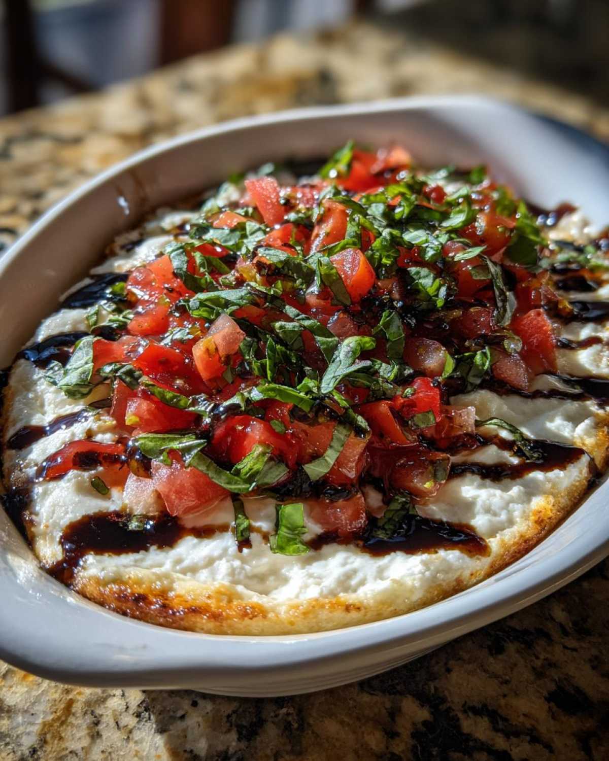 A close-up of hot baked bruschetta dip topped with fresh diced tomatoes, basil, and a balsamic glaze drizzle.