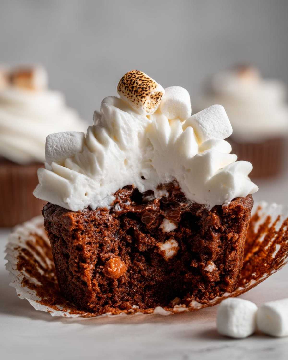 Close-up of a hot chocolate cupcake cut in half, showing rich chocolate cake, melted chocolate chips, and white frosting topped with toasted marshmallows.