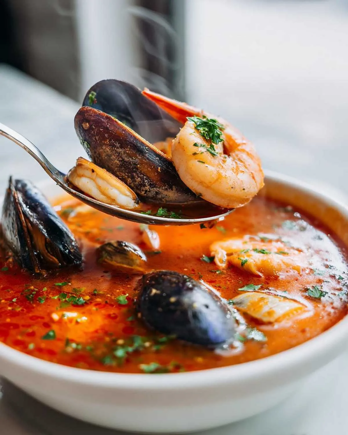 A spoonful of hot seafood soup lifts a shrimp and mussels from a bowl of rich, red seafood soup.