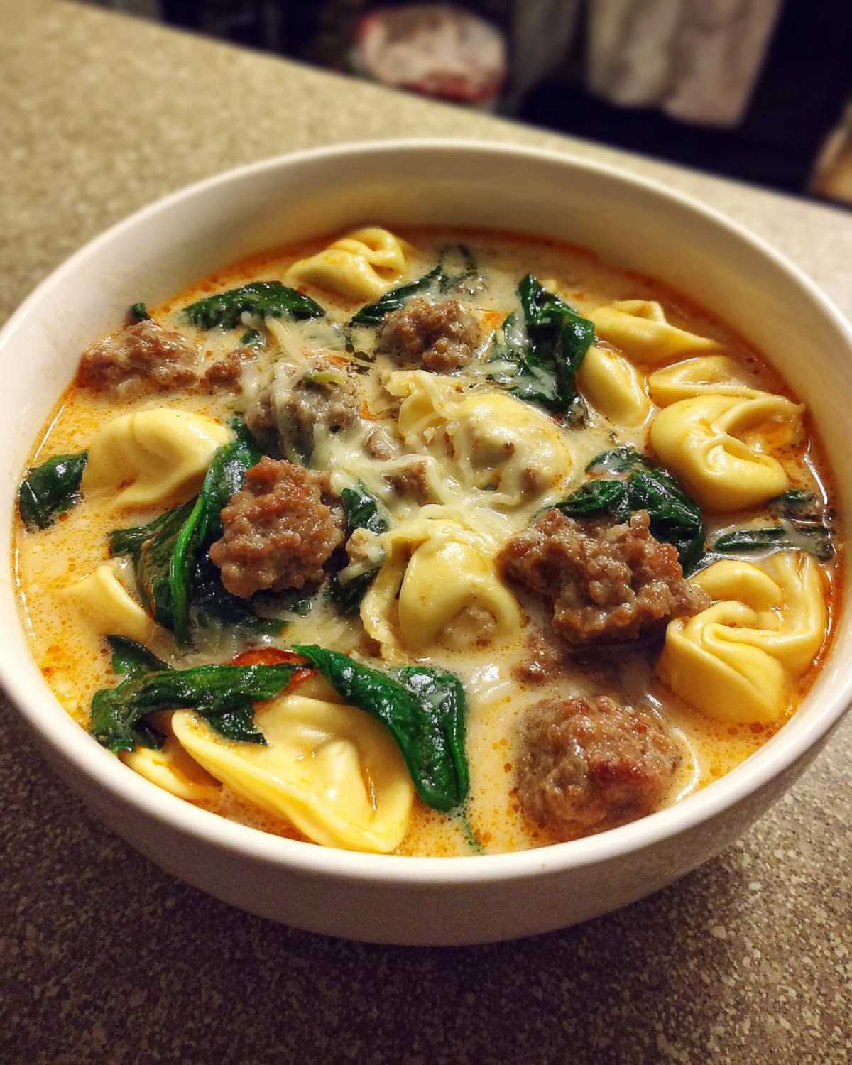 A close-up of a creamy bowl of italian sausage tortellini soup with spinach and melted cheese.