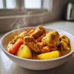 A close-up of a steaming white bowl filled with rich, yellow Jamaican curry chicken, potatoes, and carrots.