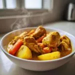 A close-up of a steaming white bowl filled with rich, yellow Jamaican curry chicken, potatoes, and carrots.