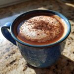 Close-up of rich Mexican hot chocolate topped with foam and cinnamon in a textured blue mug.