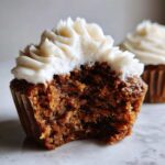 Close-up of a moist gingerbread cupcake with a bite taken out, showing the rich brown crumb and white frosting.