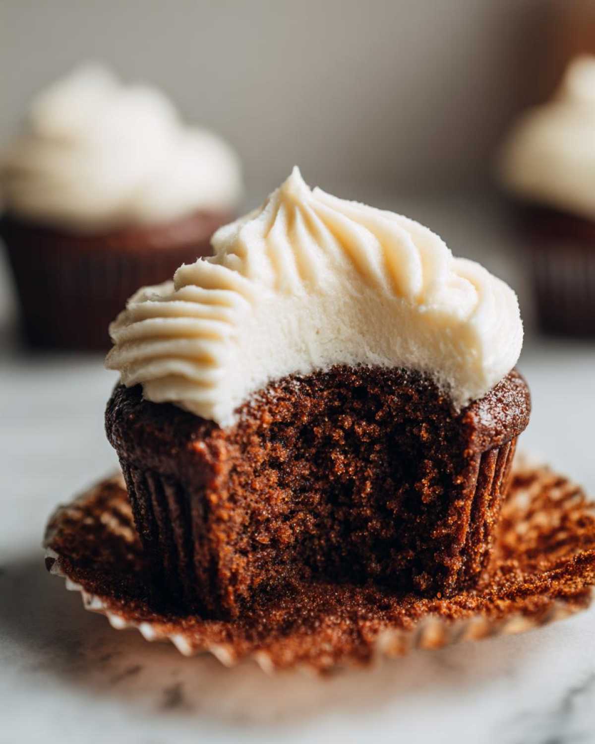 Close-up of a moist gingerbread cupcake with a bite taken out, showing the dark crumb and thick cream cheese frosting.