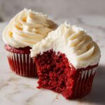Close-up of a moist red velvet cupcakes, one bitten into, showing the rich red crumb and thick cream cheese frosting.