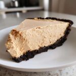 A thick slice of no bake peanut butter pie featuring creamy filling and a dark chocolate cookie crust on a white plate.