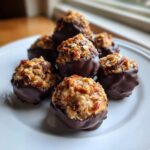 A close-up of several homemade pecan pie balls dipped in dark chocolate, resting on a white plate.