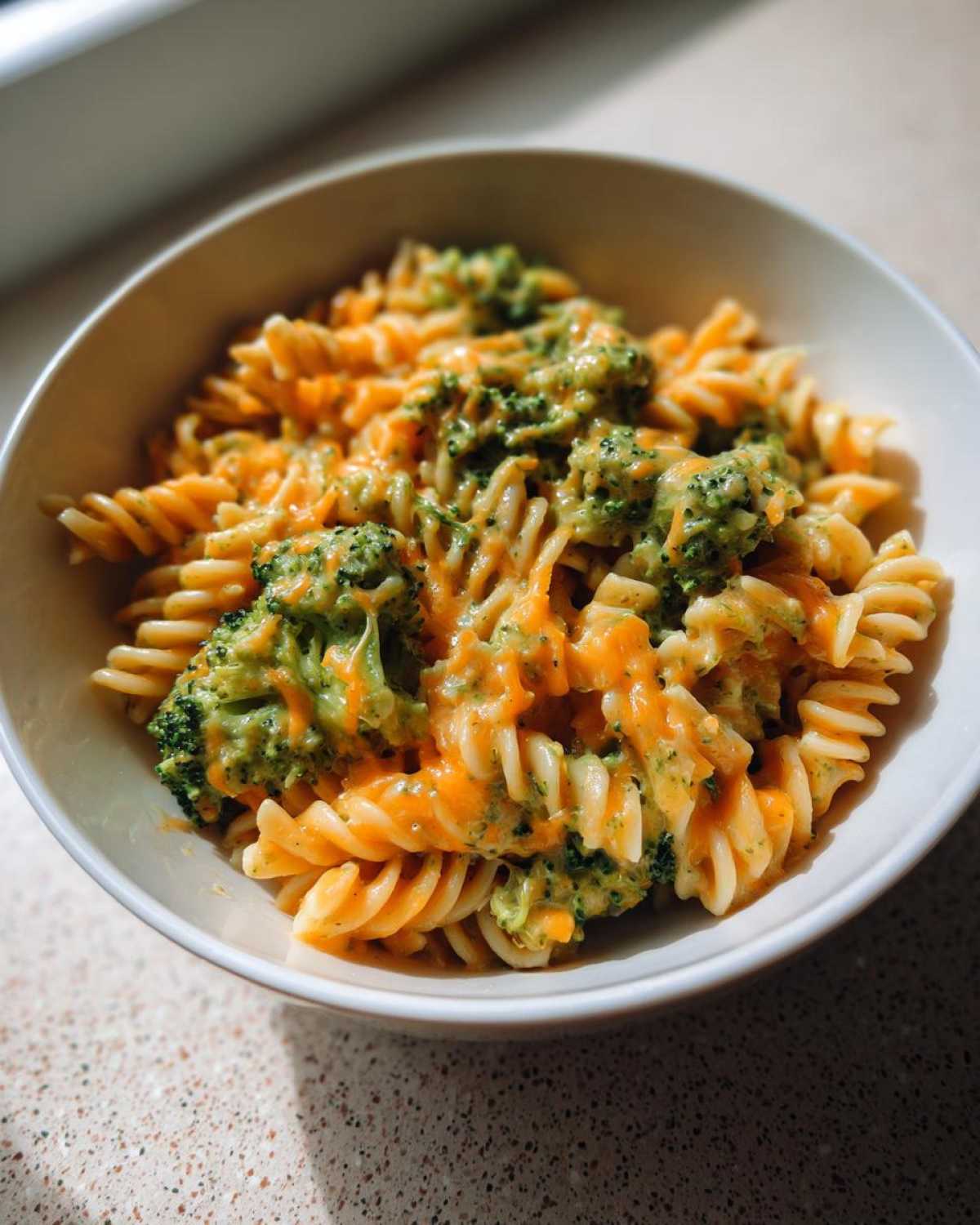 Close-up of a bowl of creamy broccoli pasta made with fusilli noodles and melted orange cheddar cheese.