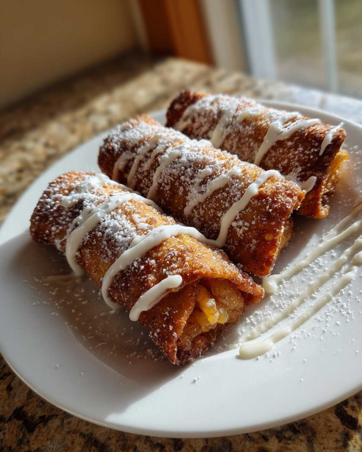 Three golden-brown peach cobbler egg rolls dusted with powdered sugar and drizzled with white icing.