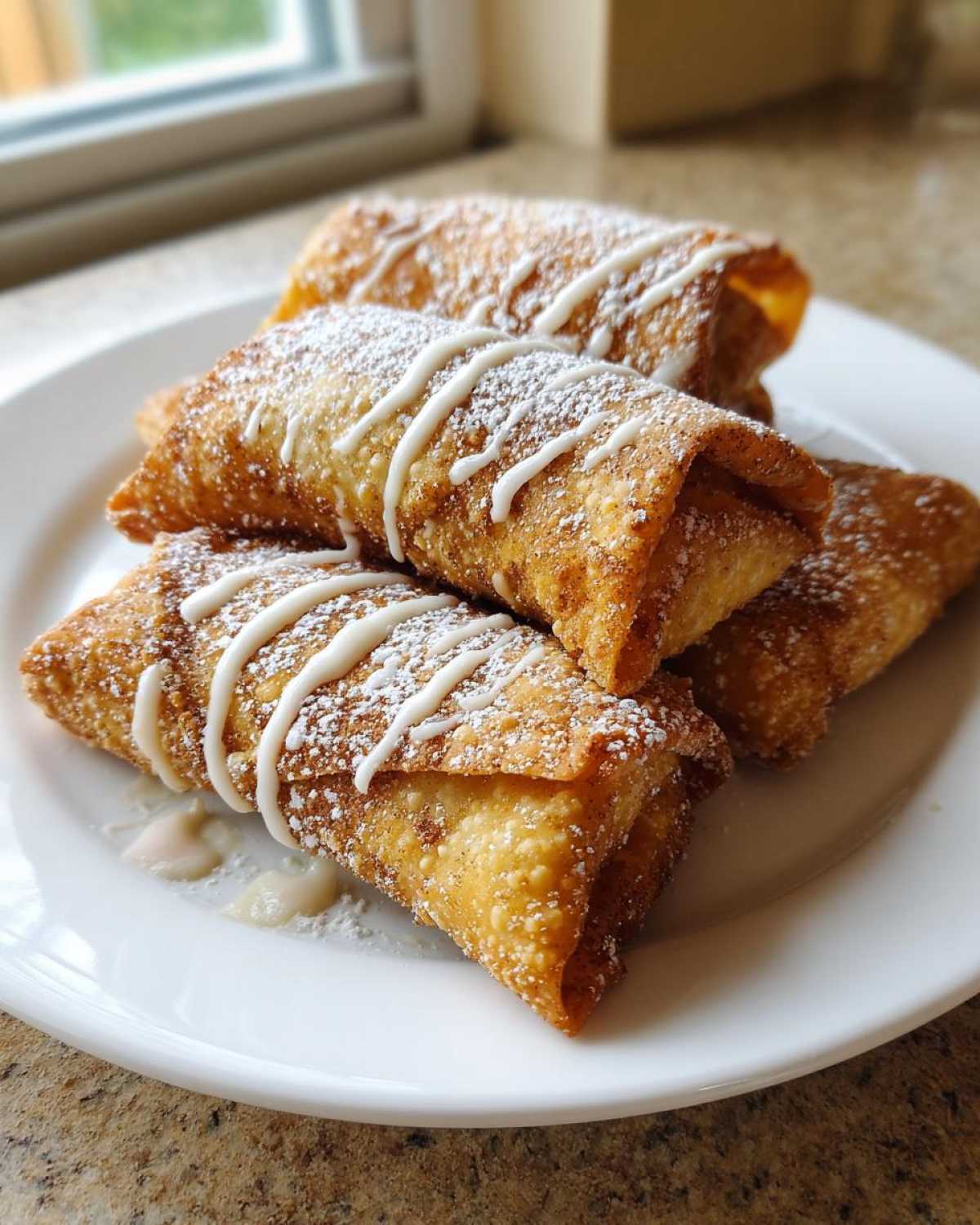 Four golden brown peach cobbler egg rolls stacked on a white plate, drizzled with white icing and dusted with powdered sugar.