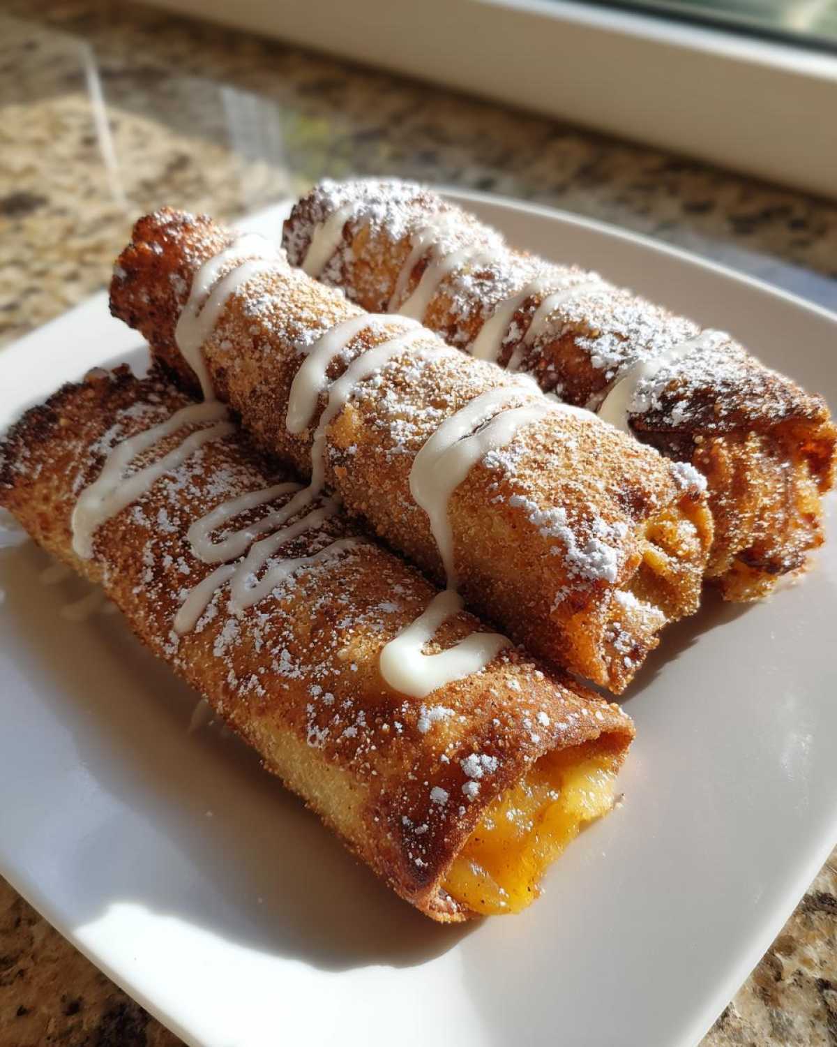 Four crispy, golden brown peach cobbler egg rolls dusted with powdered sugar and drizzled with white icing.