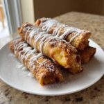 A stack of four crispy, golden peach cobbler egg rolls drizzled with white icing on a white plate.