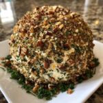 A large, round cheese ball recipe coated entirely in chopped pecans, herbs, and spices, resting on a white platter.