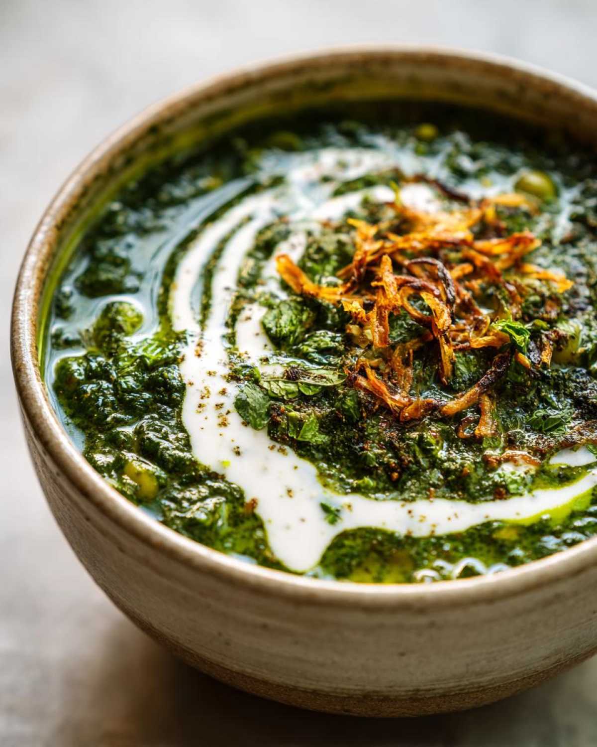 Close-up of a bowl of vibrant green Persian noodle soup, topped with yogurt swirl and crispy fried onions.